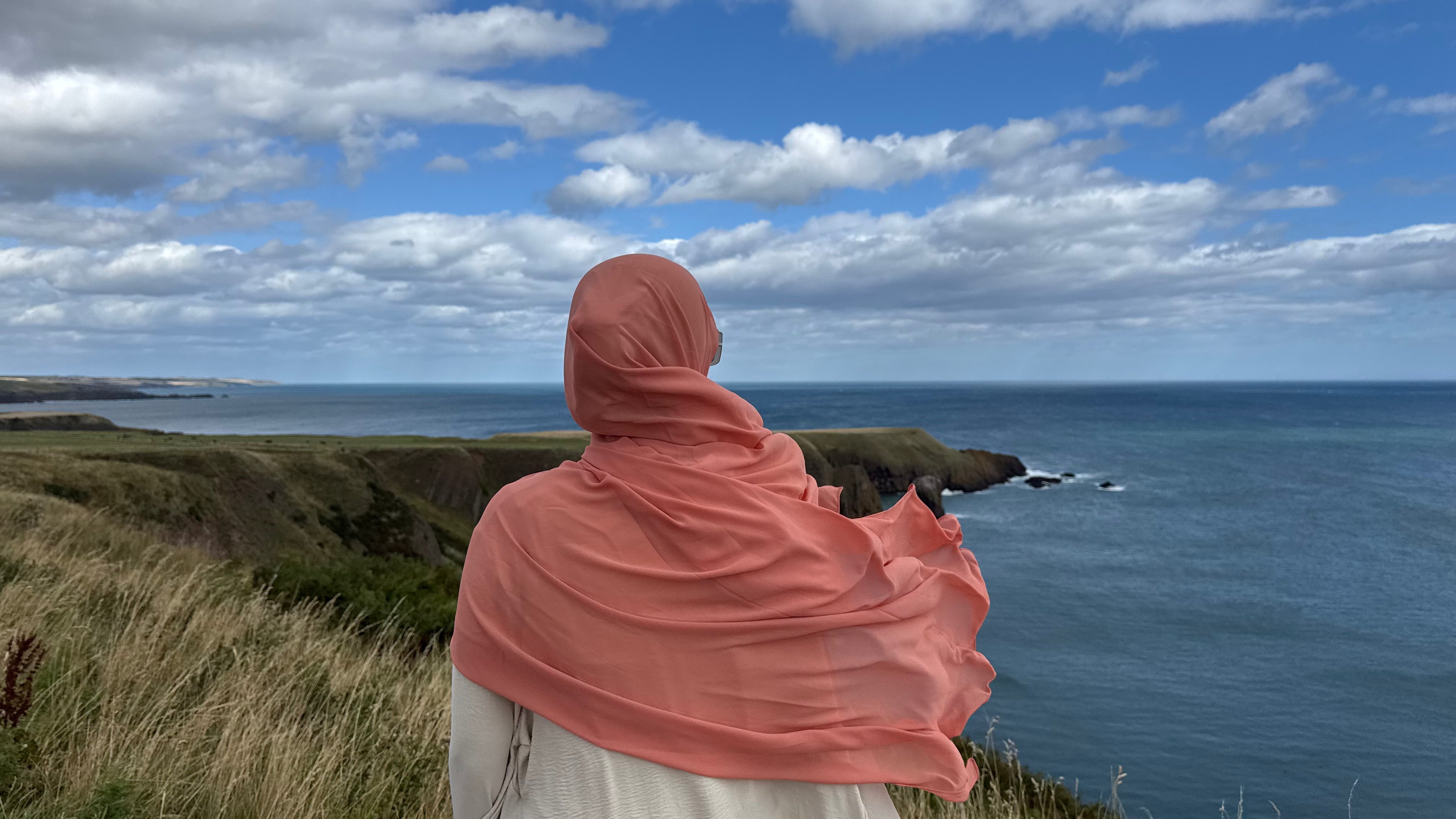 Person wearing a pink hijab overlooking a coastal landscape with blue sky and clouds.